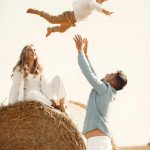 Joyful family moment catching son mid-air on hay bale in countryside.